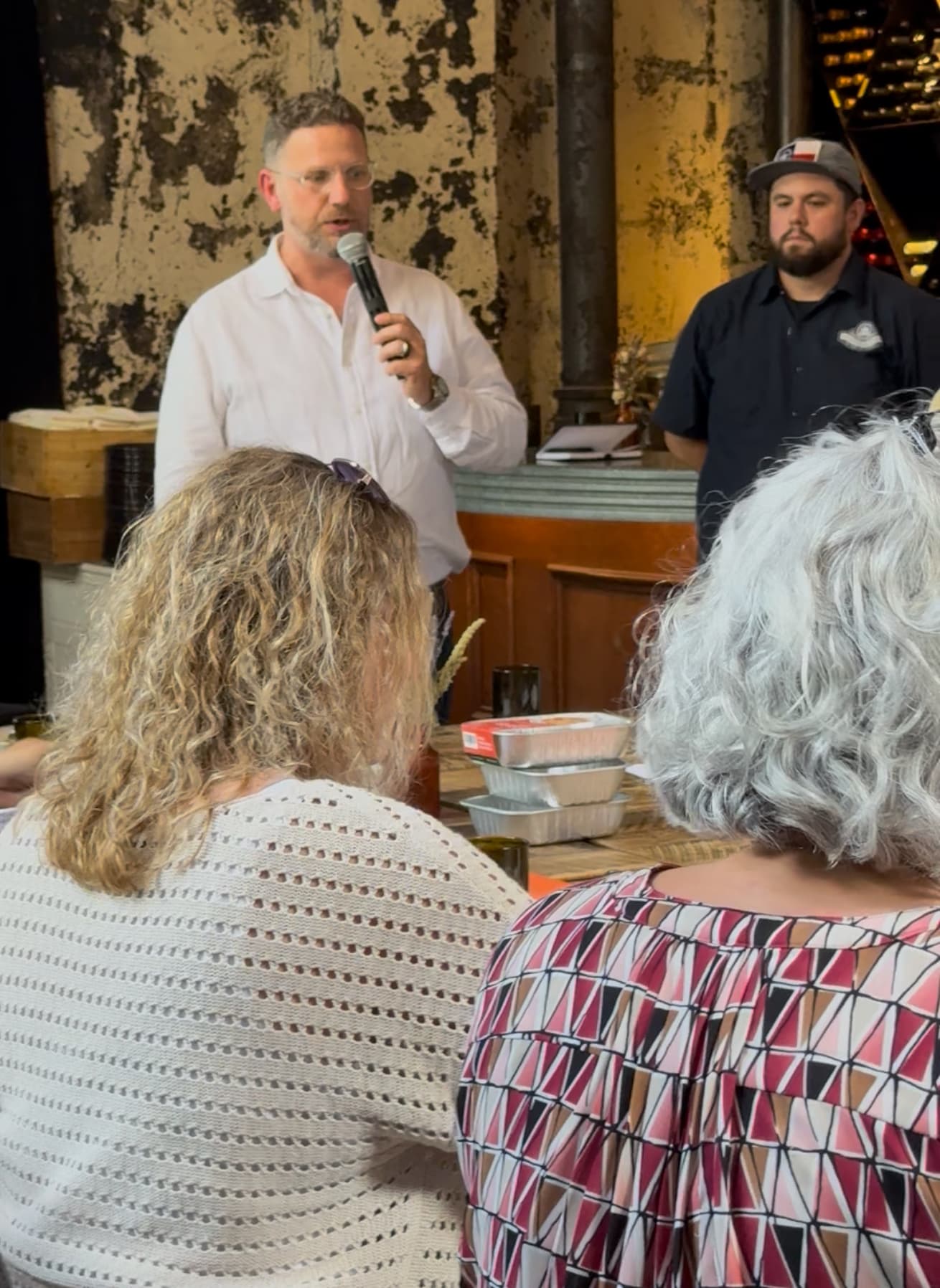 A speaker with a microphone addressing seated guests at a tasting, with foil meal trays stacked on the table